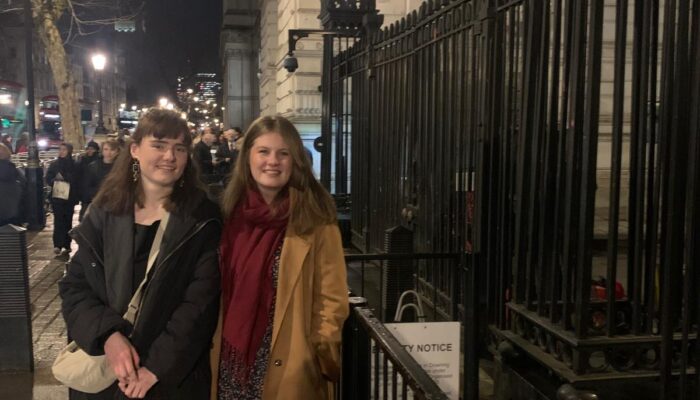 Two women standing on a London sidewalk at night, positioned next to a black iron fence near a Downing Street sign.