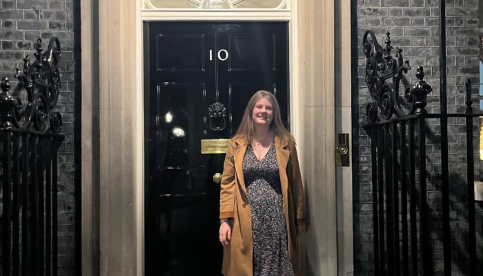 Laura Penman stands in front of the iconic black door of 10 Downing Street, illuminated by an overhead light at night.