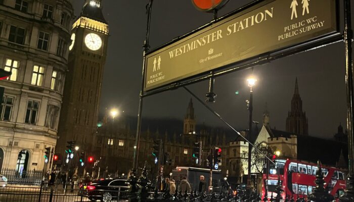 A night scene of Westminster Station’s entrance sign in London, with a red double-decker bus and a lit-up Big Ben in the background.