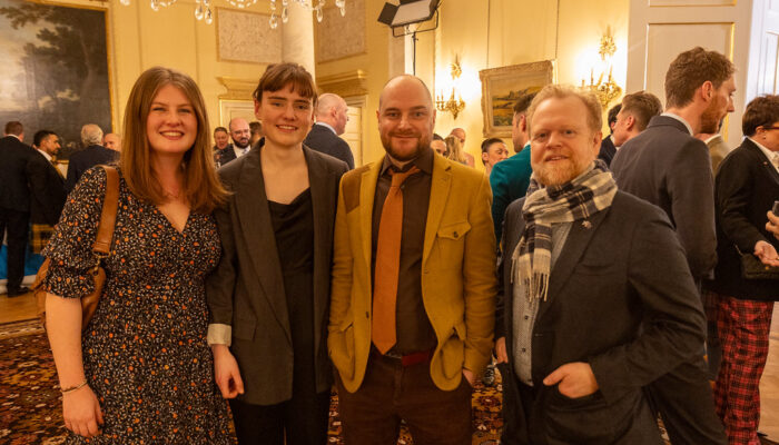 Four people posing for a photo in a brightly lit, elegant ballroom under a crystal chandelier during a formal event.