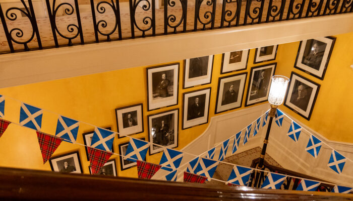 An ornate staircase featuring yellow walls decorated with several framed black-and-white portraits and blue-and-white Scottish saltire bunting.