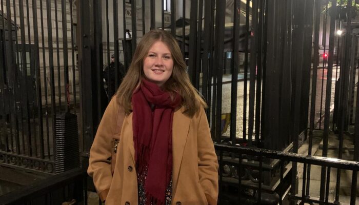 A woman in a tan coat and red scarf smiling while standing in front of a tall black iron gate at night.